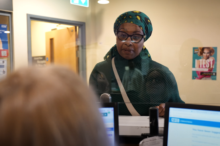 Black lady in GP reception talking to receptionist behind a clear screen
